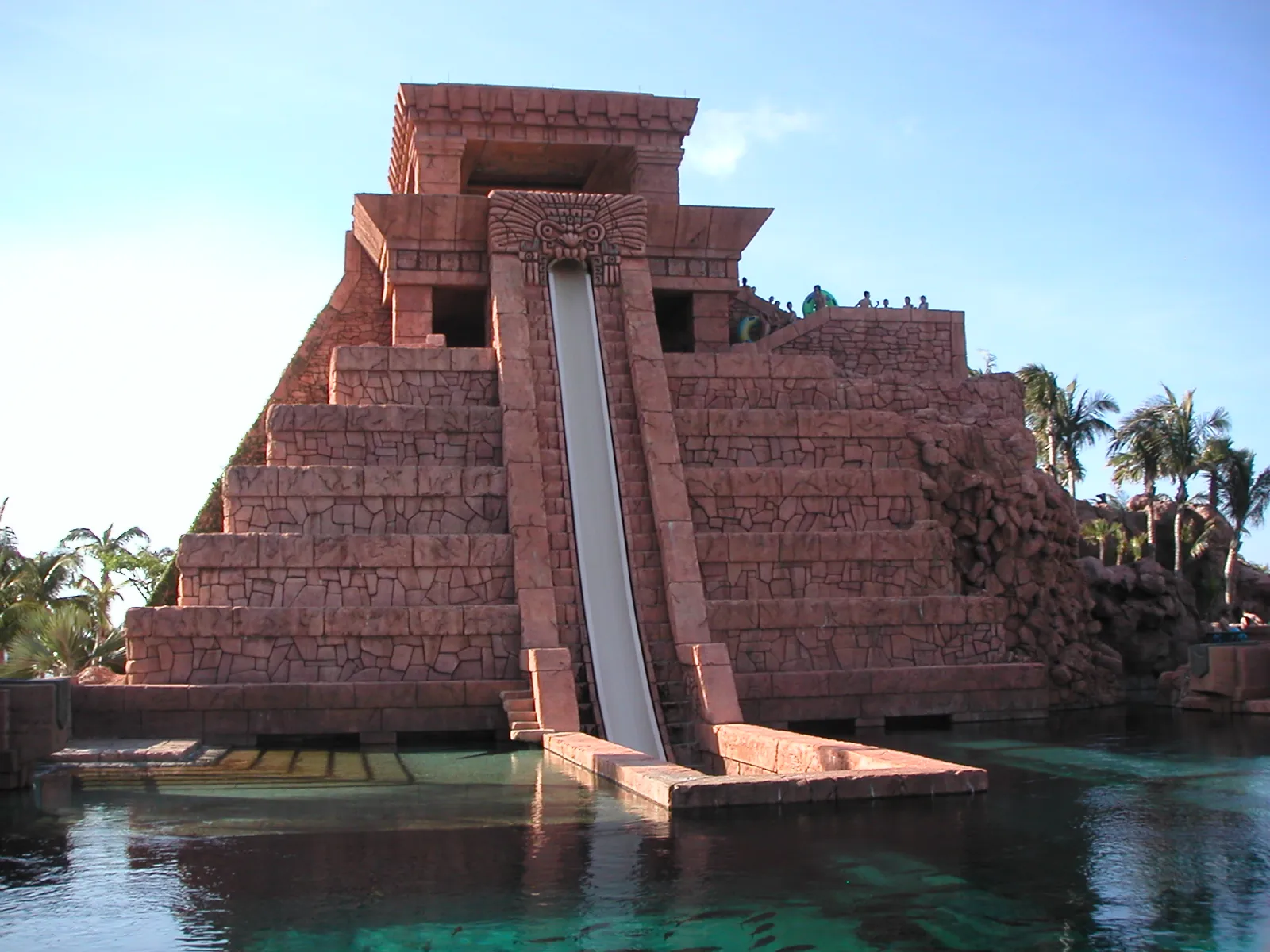 A water slide drops steeply down the front of a tall Mayan-style stone temple into a turquoise pool at Atlantis, with palm trees and people at the top