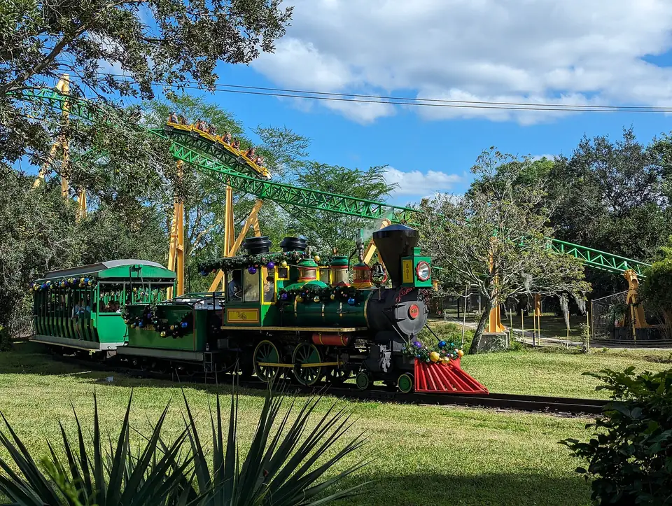 A green steam-style train decorated with holiday garlands passes a lawn at Busch Gardens Tampa Bay while riders go by on a green and yellow roller coaster behind the trees