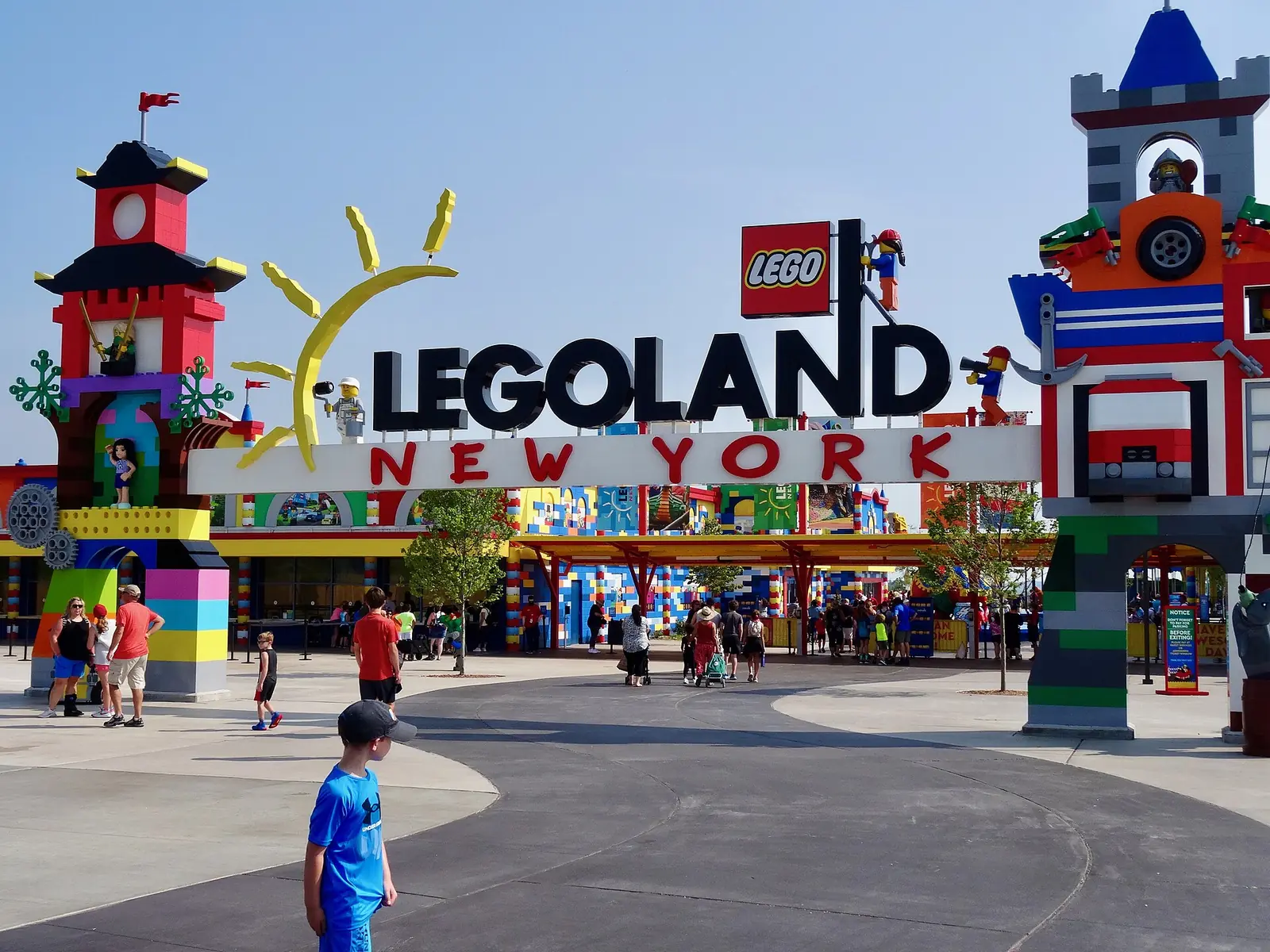 The LEGOLAND New York entrance on a sunny day: a big LEGOLAND New York sign between two colorful brick towers, with families walking toward the gates and a boy in a blue shirt in front