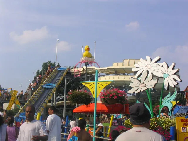 Riders carry blue rafts up a stairway to a tall yellow water slide topped by a giant rubber duck at Sesame Place, with big white flower decorations and families in the foreground
